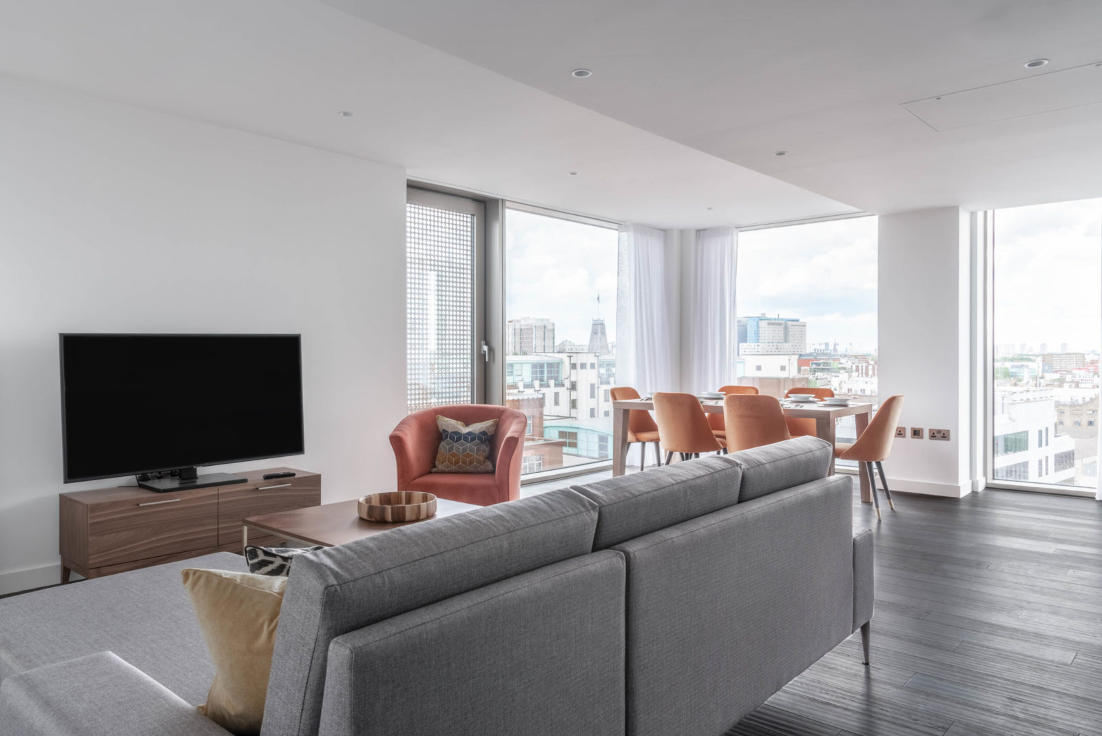Sitting area with sofa, individual chairs, a tv and a dining table with chairs in the background. Room has large floor to ceiling windows with views of London.
